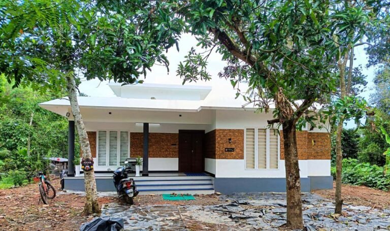 Exterior view of Ravindram, a 1800 sq. ft. modern house in Puthenchira, Thrissur, featuring a white facade with texture work, a spacious sit-out with matte finish tiles and granite, and steel-framed windows, designed for elegance and functionality.